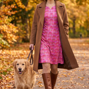 Eine Frau trägt ein Herbstkleid in pinkem Muster mit einem Mantel und Stiefeln in Braun. Sie führt einen Hund an der Leine.